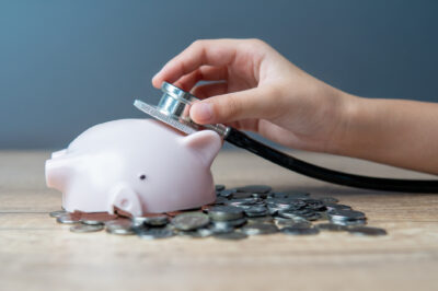A hand holding a stethoscope listening to a broken piggy bank lying on a pile of coins.